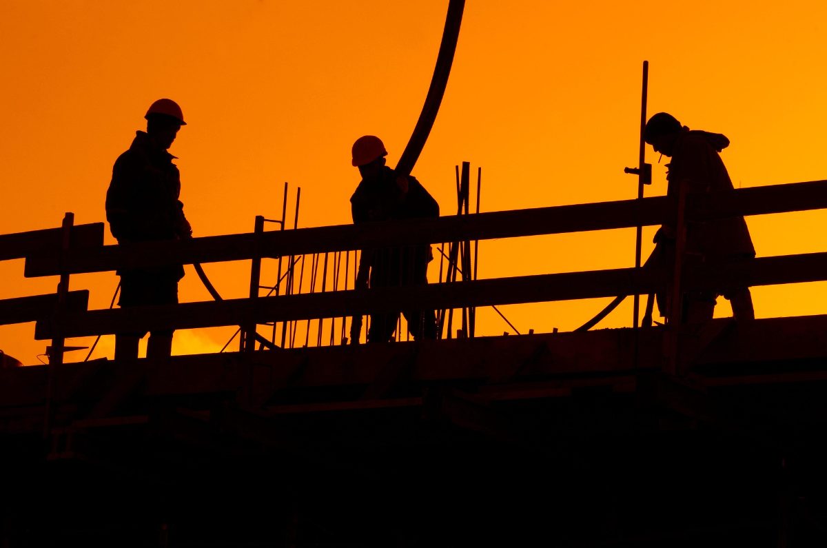 Bauarbeiter mit Helmen als Silhouette vor orangem Abendhimmel auf einem Gerüst